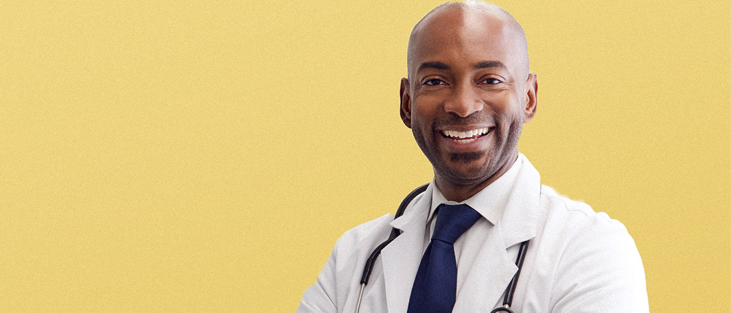 A middle-aged Black male doctor in a white labcoat and navy tie smiling against a yellow background.