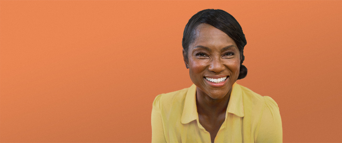A middle-aged Black woman in a yellow shirt smiling against an orange background.